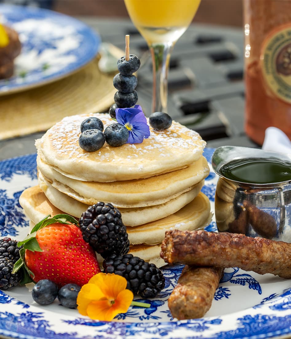 Stack of pancakes with blueberries, fresh fruits, sausage, and syrup