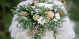 A bride holding a snow-dusted winter wedding bouquet of roses, pine needles, and pine cones