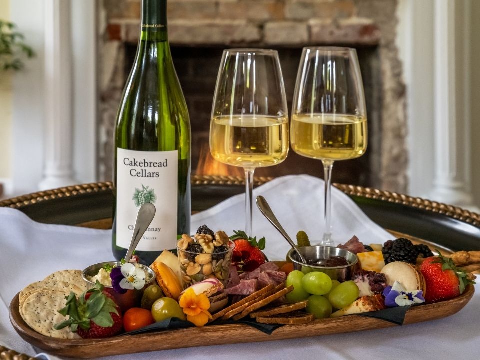 A wooden platter of meats, cheeses, and fruit in front of a bottle of wine and two full wine glasses